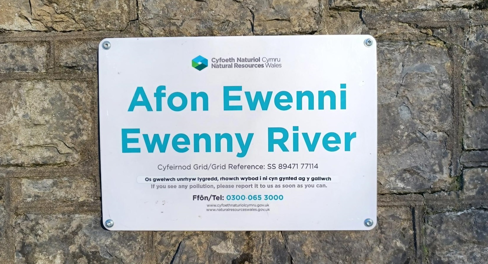Arwydd ar wal wrth ymyl afon gyda gwybodaeth ar sut i roi gwybod am lygredd